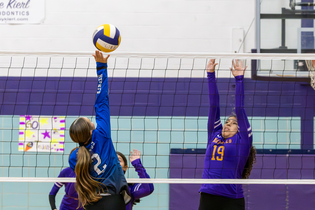 A Hefner player jumps with both arms extended at the net as Mayfield player number 28 prepares to block.