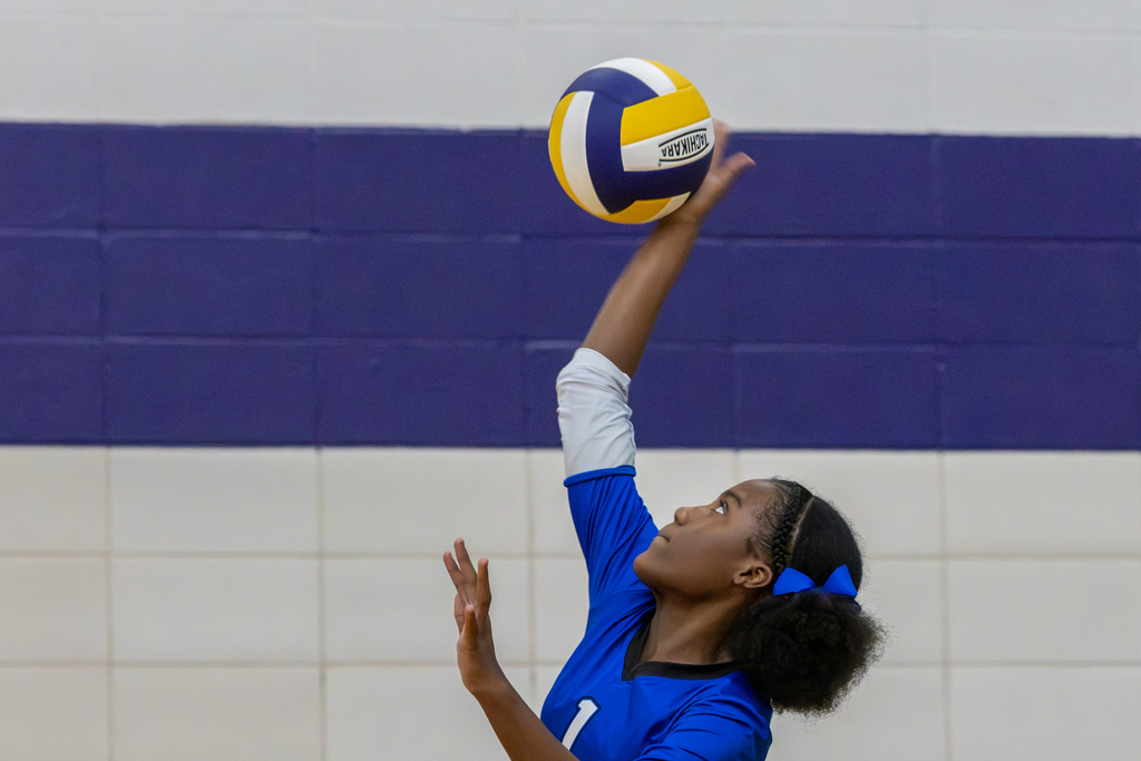 A Hefner volleyball player reaches high to serve the ball, looking up toward it as she begins the play.