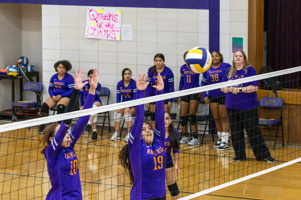 Mayfield players number 10 and 19 jump with their hands raised to block the ball at the net while teammates watch from the bench.