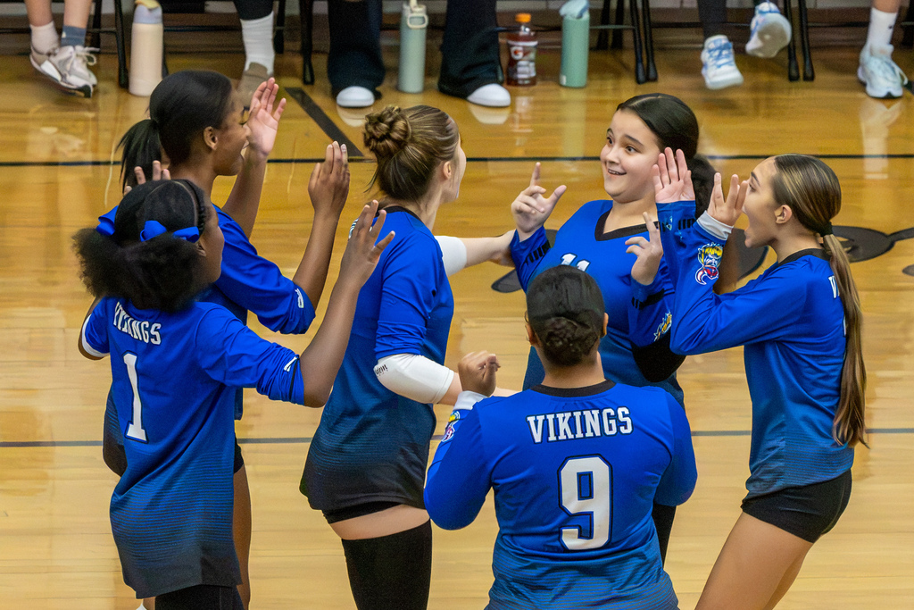 Hefner volleyball players gather in a circle, smiling and raising their hands in celebration.
