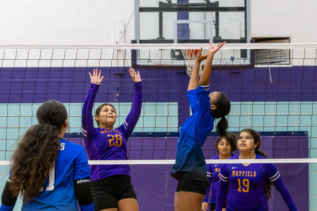 A Hefner player jumps with both arms extended at the net as Mayfield player number 28 prepares to block.
