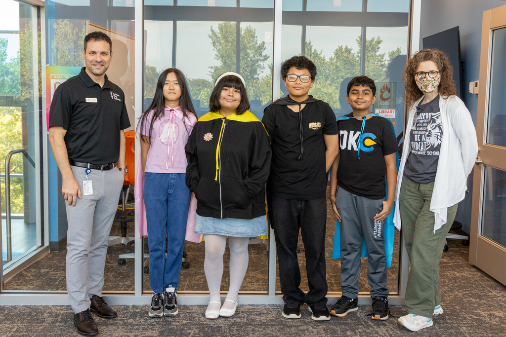 Four Capps Middle School Tech Crew students stand with two Putnam City Schools staff members in the school library. Several students are wearing colorful capes as part of their Tech Crew activity.