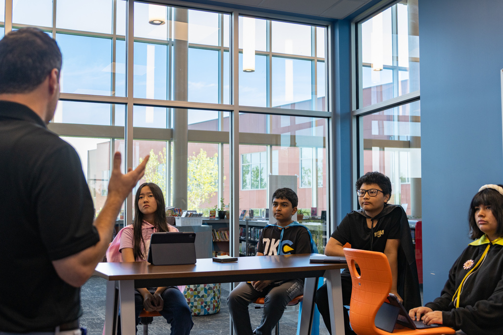 A Capps Middle School Tech Crew class listens as a Putnam City Schools Communications team member speaks about photography, videography and interviewing. Four students sit around a table with tablets in front of them.