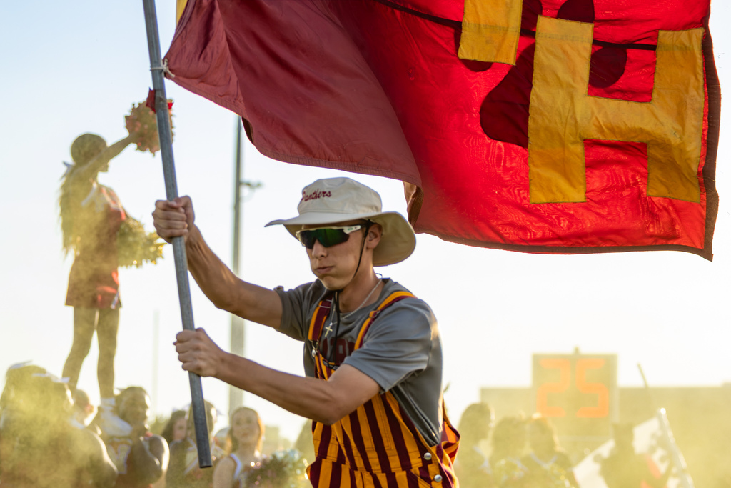 A PC North student carries a large red-and-gold flag through golden backlight as a cheerleader raises pom-poms in the background.