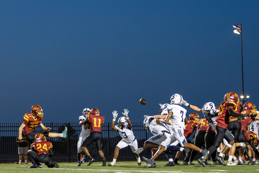 A PC North kicker sends the football over the line of scrimmage beneath the stadium lights and dark blue evening sky.