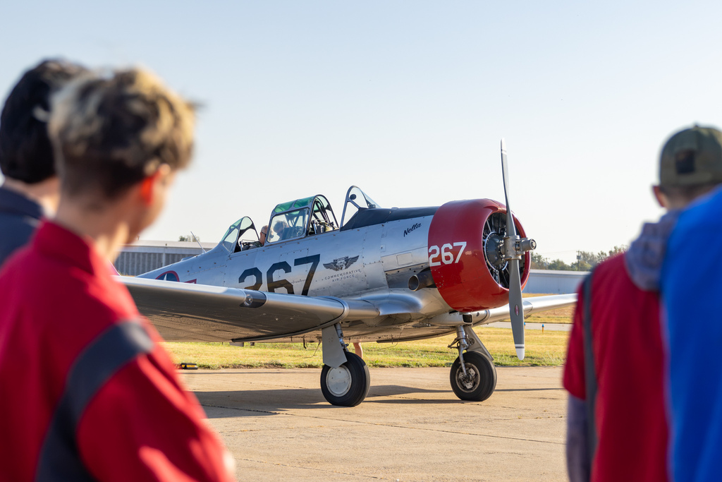 A vintage propeller aircraft sits on the ramp, framed by aviation students watching from the foreground.