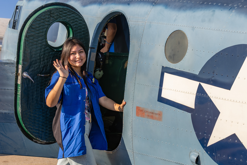 An aviation student smiles and waves from the doorway of a historic aircraft.