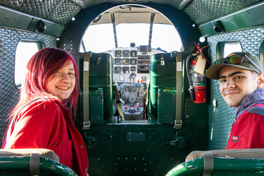 Two aviation students smile inside a historic aircraft with the cockpit visible behind them.