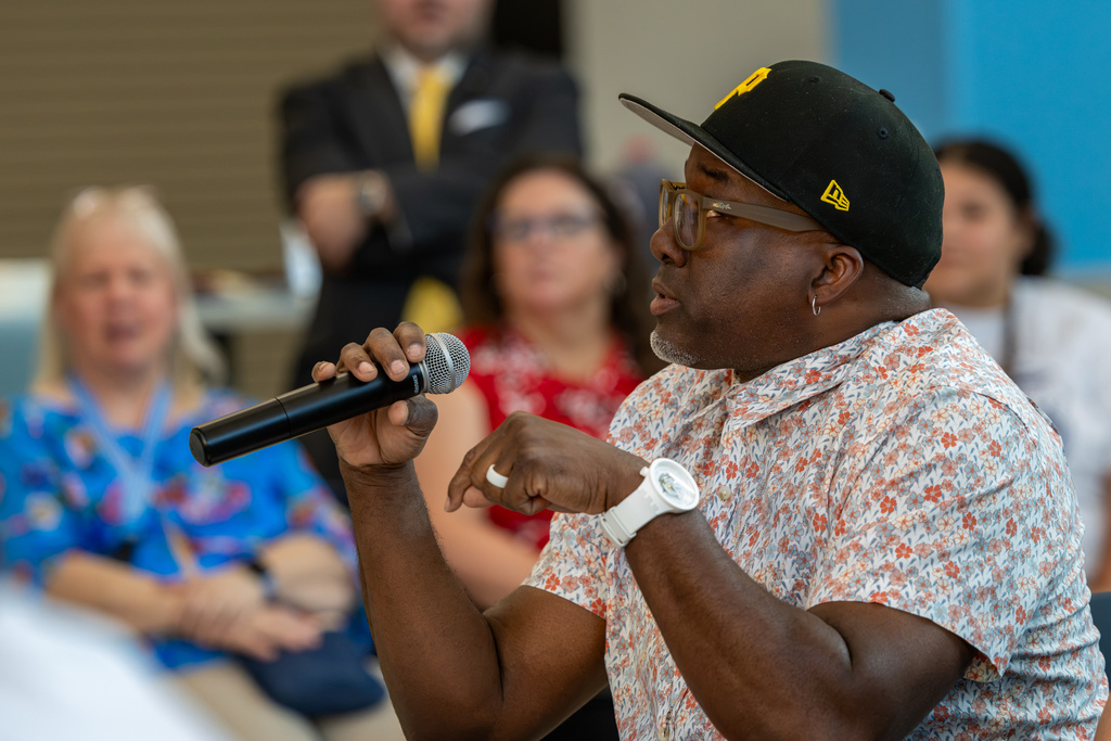 A man in a floral shirt and black cap speaks into a microphone while seated among other Real Talk attendees.