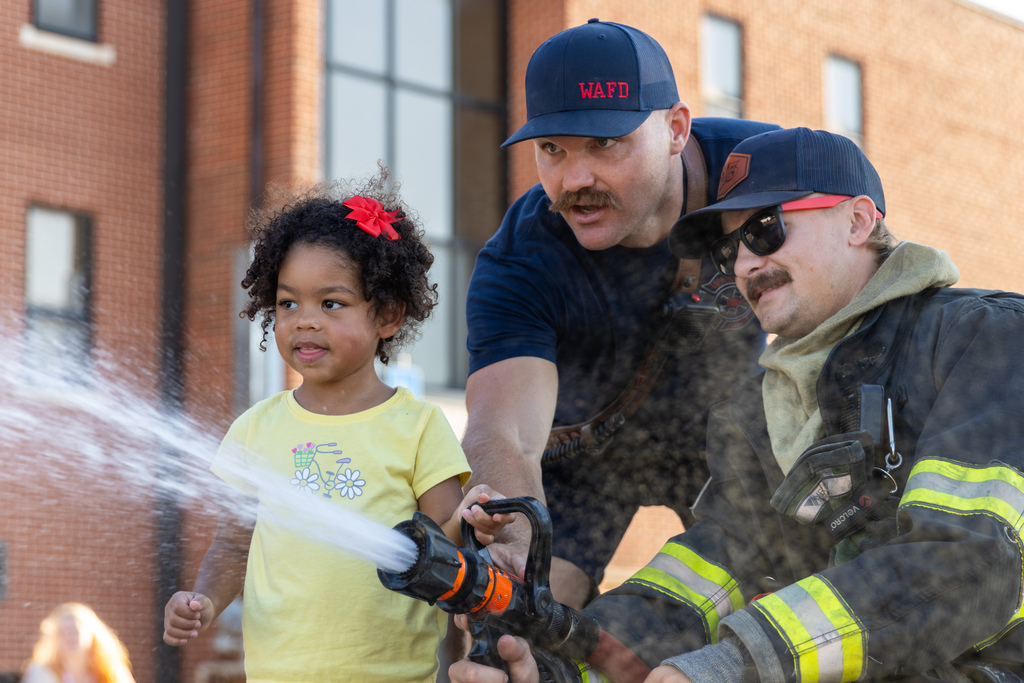 A Smart Start student in a yellow shirt sprays water from a fire hose with help from two Warr Acres firefighters.
