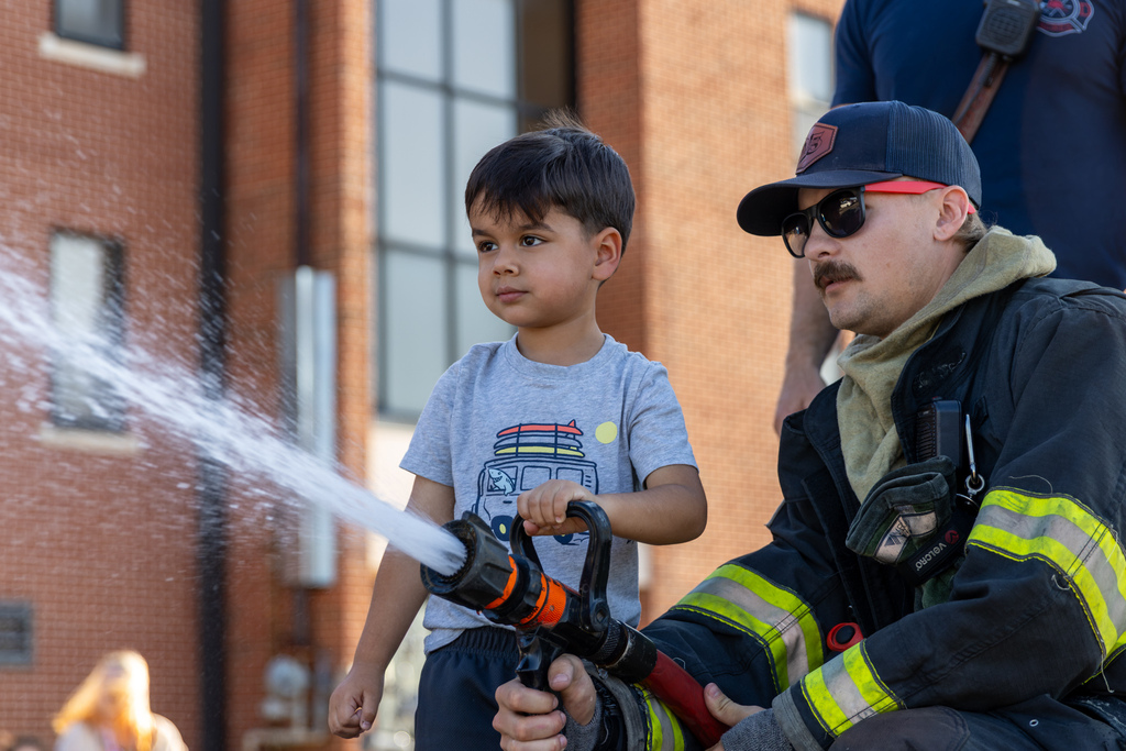 A Smart Start student holds a fire-hose nozzle as a firefighter in protective gear helps direct the stream of water.