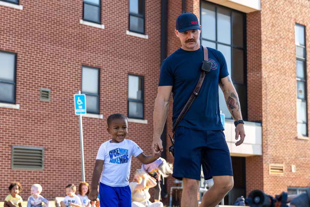 A smiling Smart Start student holds a Warr Acres firefighter’s hand after taking his turn with the hose.