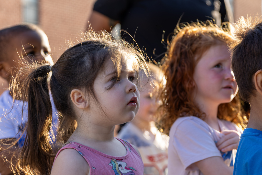 A Smart Start student in a pink unicorn shirt watches a firefighter’s demonstration while classmates gather around her.