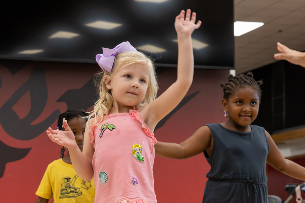 A young dancer in a pink dress and purple bow raises one arm while practicing choreography, with other children dancing behind her in the PC North Commons.
