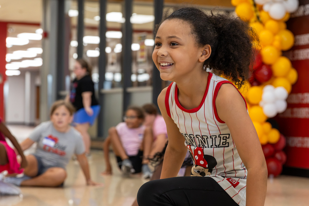 A young dancer smiles broadly while crouched during practice, with other participants seated behind her near red and gold balloons.