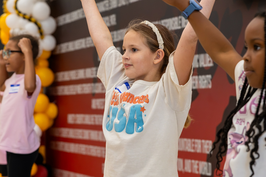 A young dancer in a white shirt raises both arms during the routine while other participants practice beside her.
