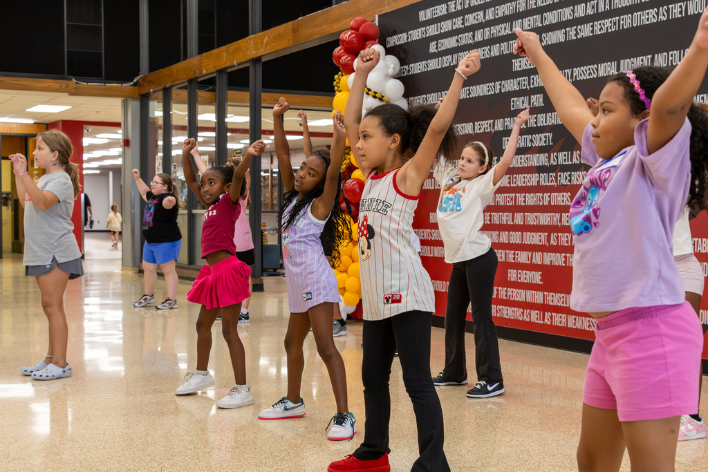 A group of young dancers practices in formation, lifting both arms overhead as they follow choreography in the PC North Commons.