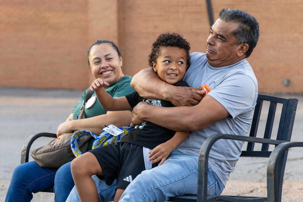 A smiling family sits together on a bench outside DD Kirkland, with a young student leaning into an adult’s arms.