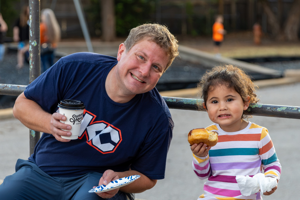 An adult and a young student sit together near the playground, holding coffee and pastries during Goodies with Grands.