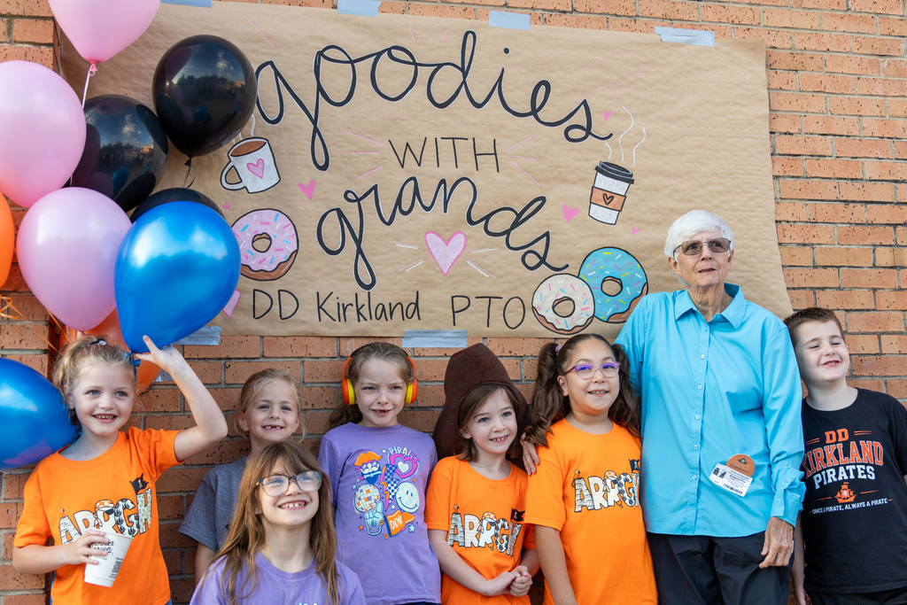 DD Kirkland students gather with an older family member beneath a handmade “Goodies with Grands” sign decorated with balloons and drawings of donuts and coffee.