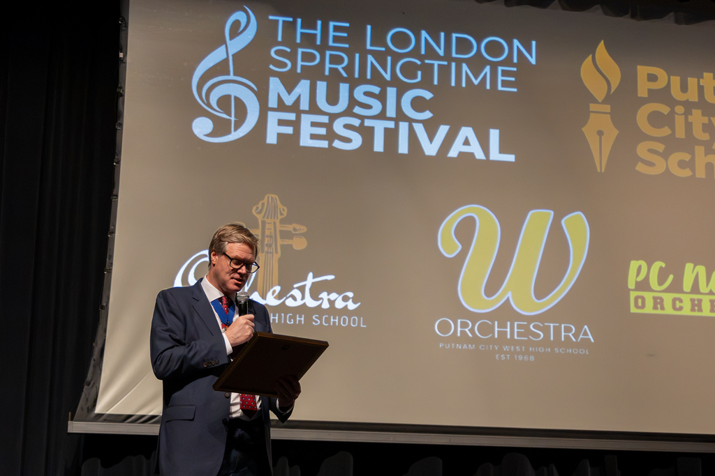 A London Springtime Music Festival representative reads from a framed presentation on stage, with the festival logo and the three Putnam City high school orchestra logos projected behind him.