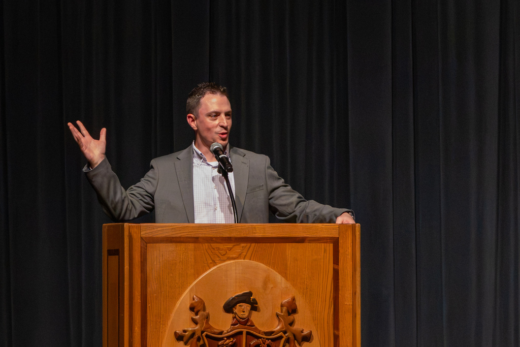 PCN Orchestra Director Jeremy Scott addresses students from a wooden podium on stage during the London Springtime Music Festival announcement.