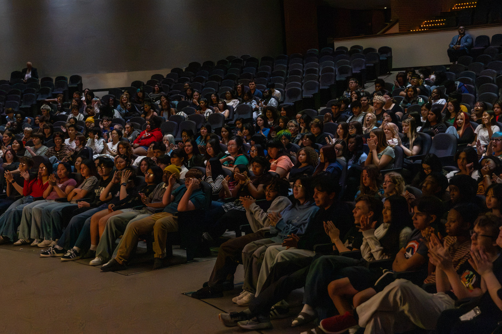 High school orchestra students fill the auditorium seats, clapping and reacting to the announcement that they have been invited to perform in London in 2028.