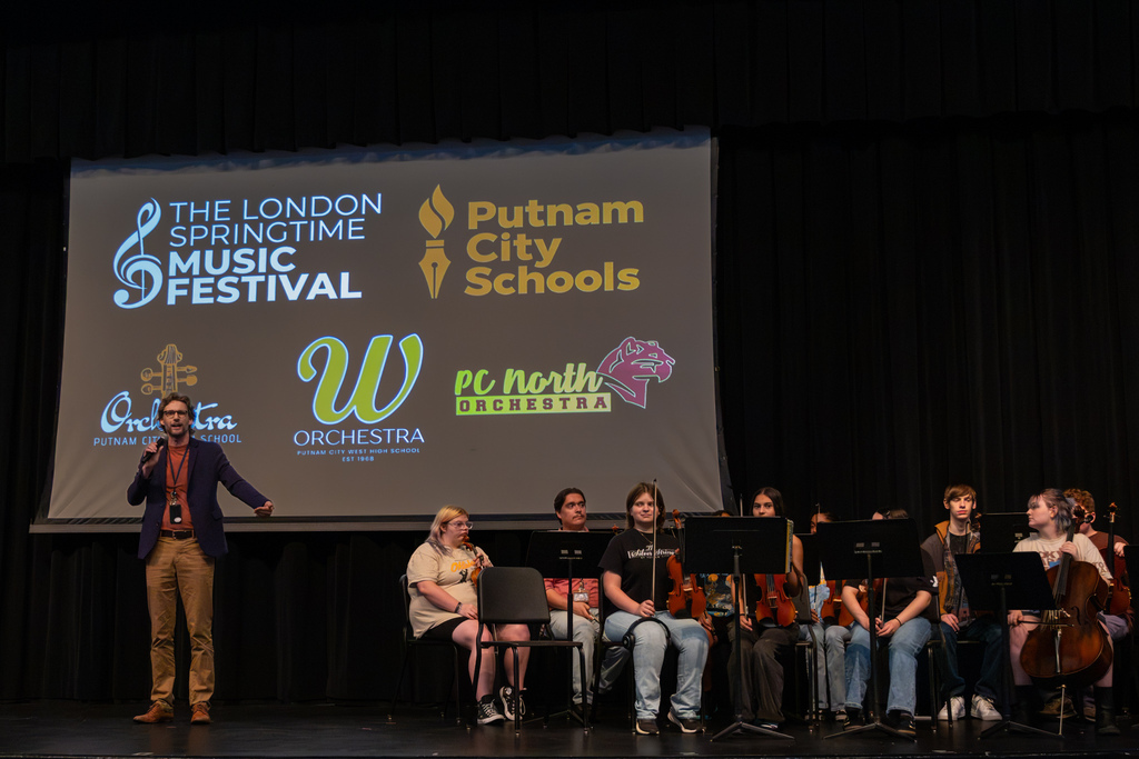 An orchestra director speaks beside seated student musicians on stage, with the London Springtime Music Festival, Putnam City Schools, and all three high school orchestra logos projected behind them.