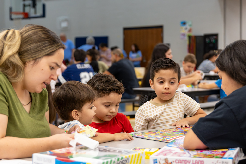 Families and students sit around tables playing board games during Central Elementary’s Family Game Night. In the foreground, several young students gather around a colorful game board while adults help them play.