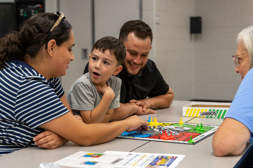 A young student reacts while playing Sorry! with family members at Central Elementary’s Family Game Night. His family smiles and watches as game pieces fill the board.