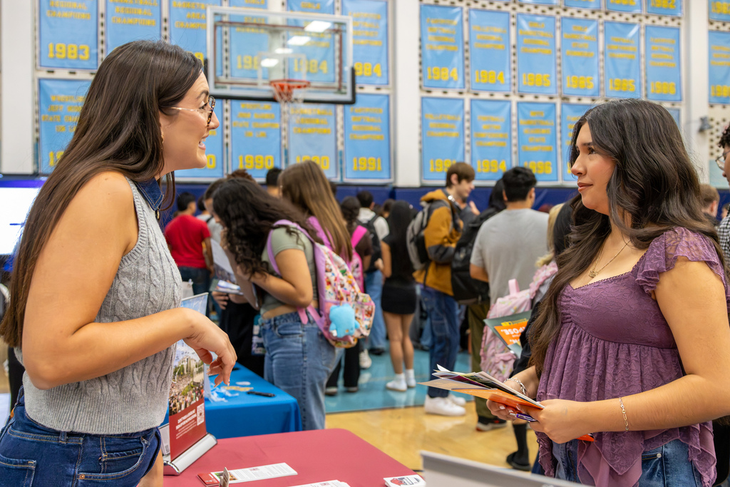 A student holding several college brochures listens as a college representative speaks with her across a table.