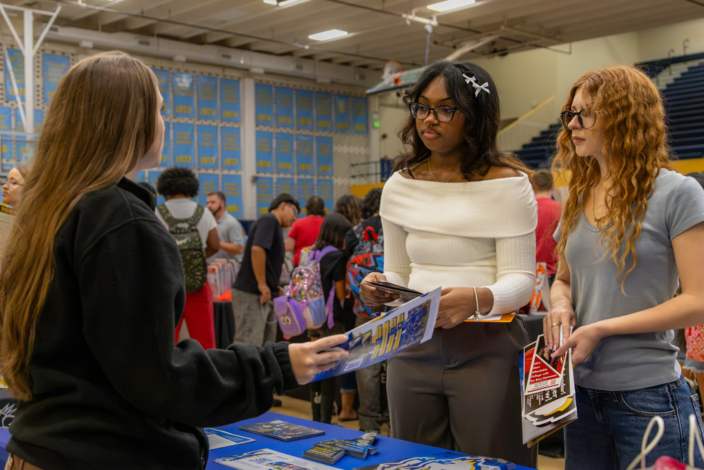 Two students holding brochures listen as a college representative shares information with them at a booth.