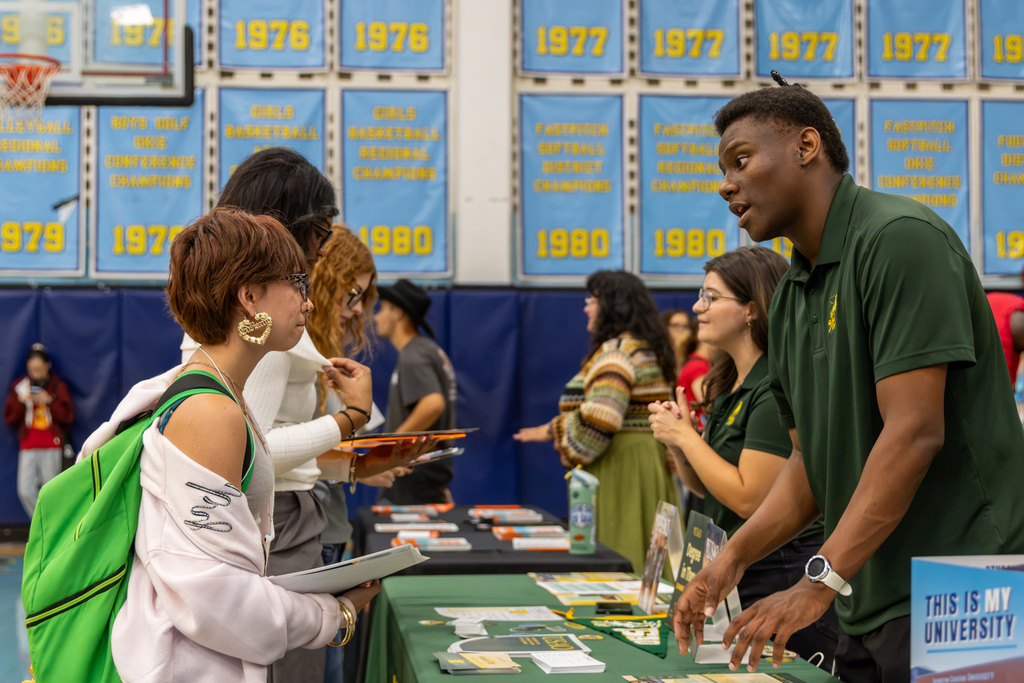 A university representative in a green polo talks with a student carrying a bright green backpack at the college fair.