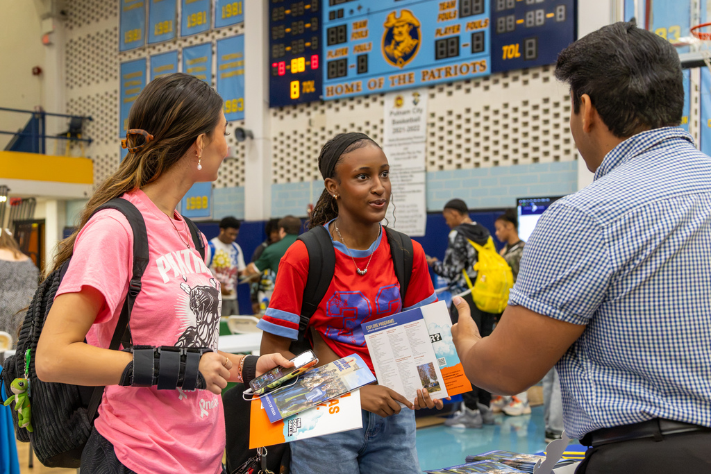 Two students holding college information talk with a representative at a booth while other students visit tables in the background.