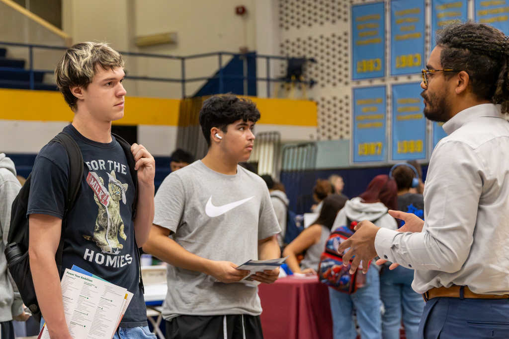 Two students listen as a college representative speaks with them at a booth inside the PC West gym.