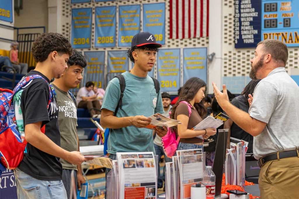 Three students holding college brochures talk with a representative at one of the college fair tables.
