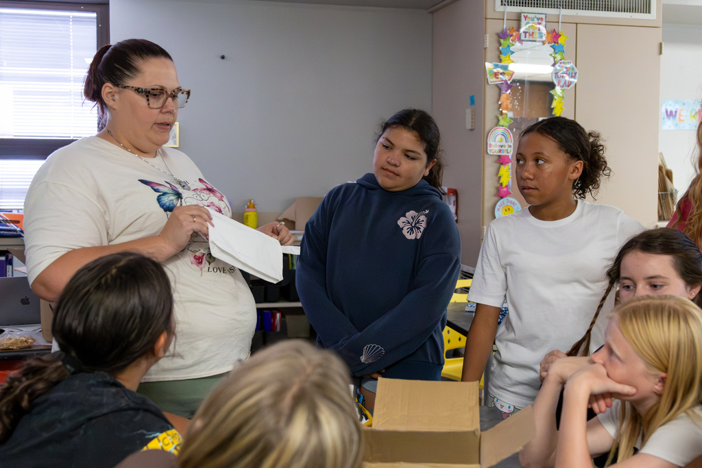 A Western Oaks Middle School coach holds up an empty hygiene bag while explaining the packing process to softball players gathered around a classroom table.