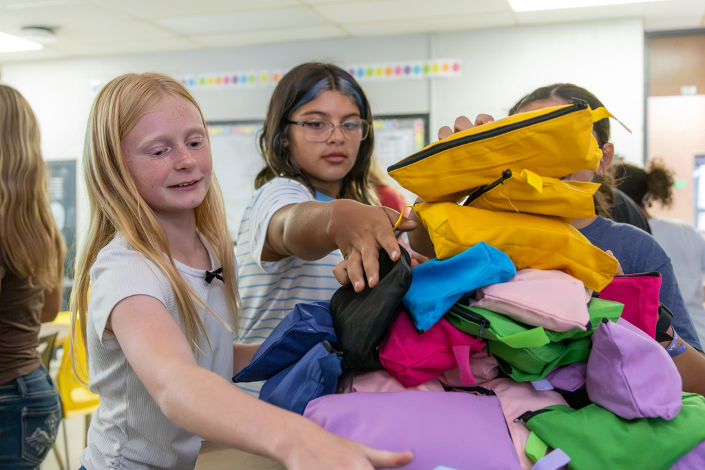 Two Western Oaks Middle School students reach toward a large pile of colorful completed hygiene bags stacked on a classroom table.