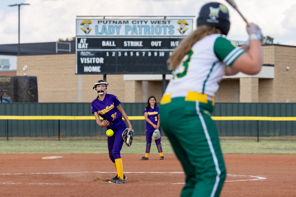 A Mayfield pitcher delivers a pitch toward home plate as a Western Oaks batter prepares to swing in the foreground.