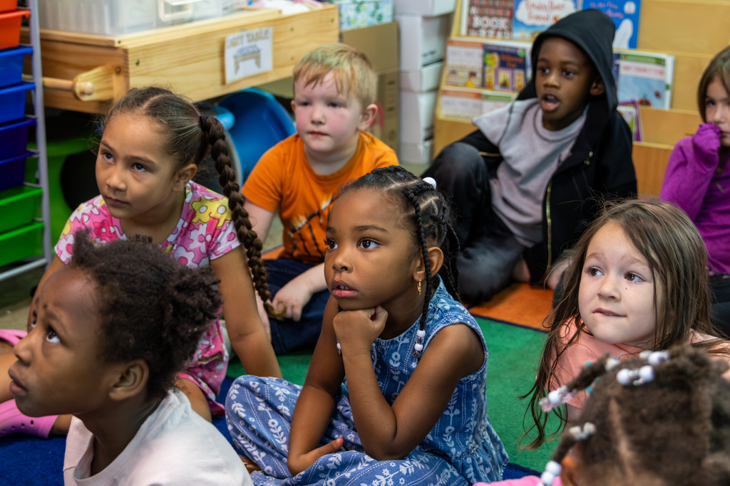 Kindergarten students sit together on a classroom rug, looking toward their teacher during a Johnny Appleseed lesson.