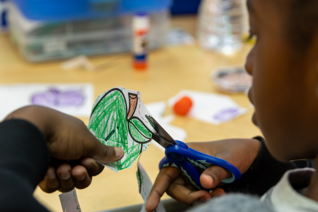 Close-up of a kindergarten student using scissors to cut out a green-colored paper piece for a Johnny Appleseed craft.