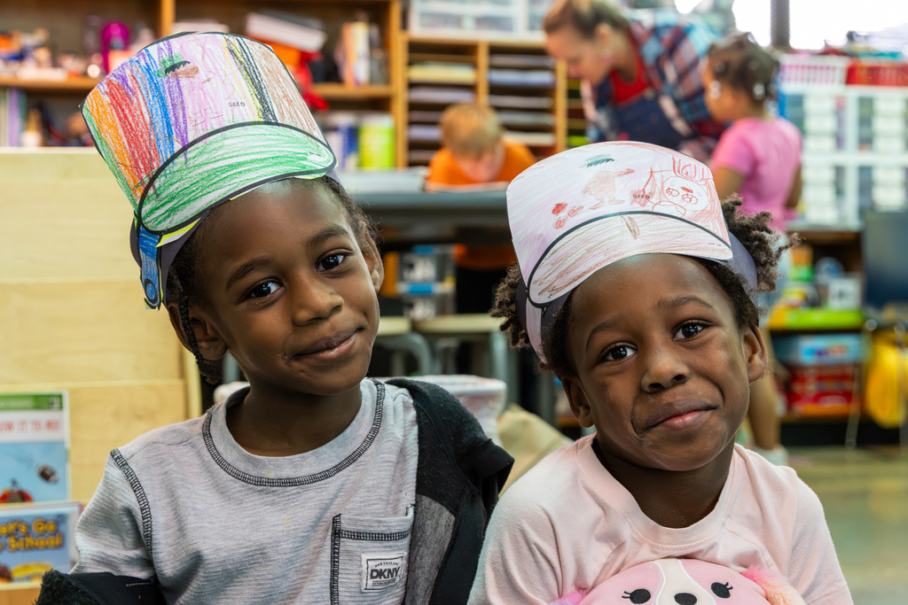 Two kindergarten students smile at the camera while wearing paper “pan hats” they colored and decorated for Johnny Appleseed’s birthday.