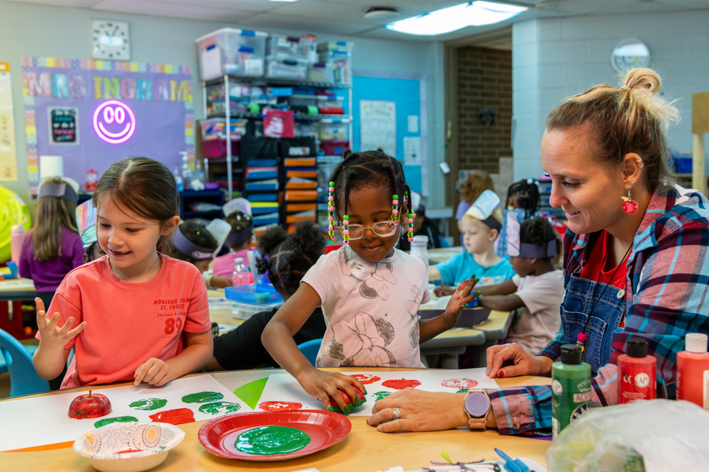 Mrs. Ingram helps two kindergarten students make red and green prints by pressing cut apples into paint and onto paper.