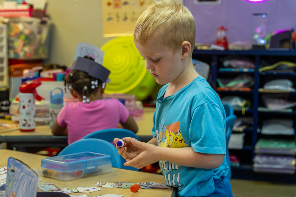 A kindergarten student stands beside a classroom table, working carefully with a glue stick during the Johnny Appleseed activity.