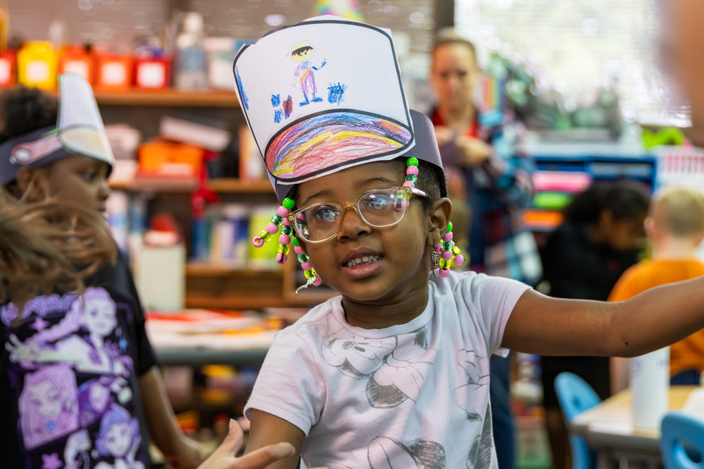 A kindergarten student wearing glasses and colorful beads smiles while showing off her decorated paper pan hat.
