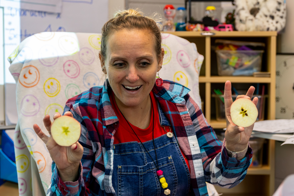 Mrs. Ingram smiles while holding up two apple halves, showing students the star-shaped pattern formed by the seeds inside.