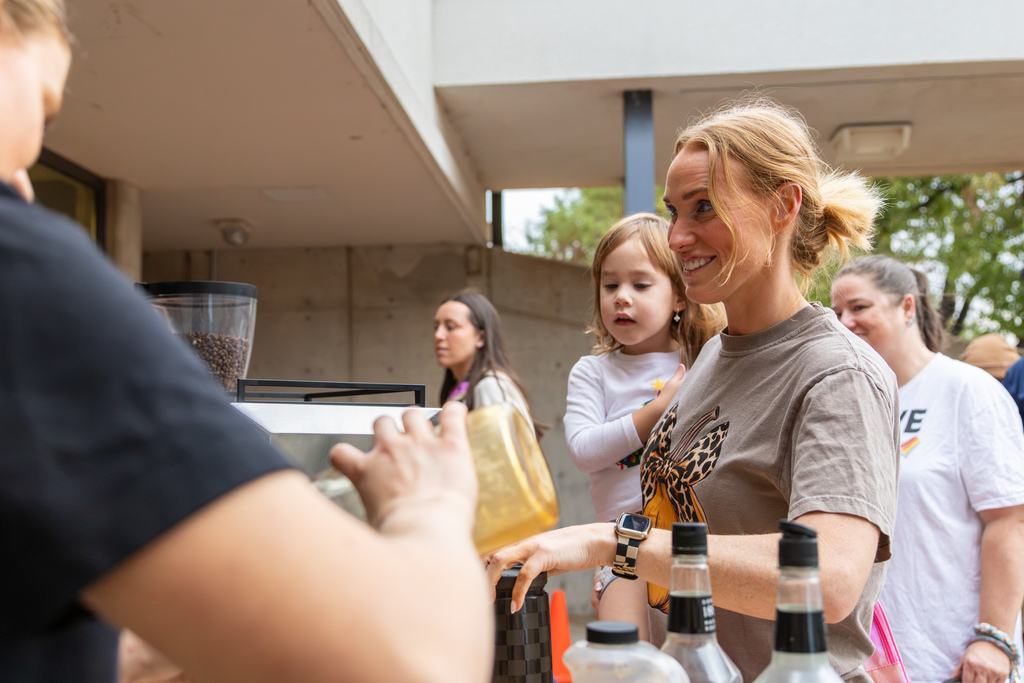 A mother holding her young daughter smiles while receiving coffee from a vendor at the outdoor coffee station.