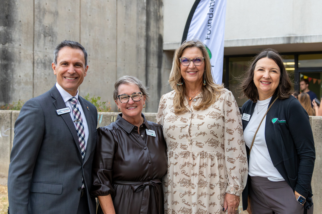 Four smiling adults pose together outside the school during the National Coffee Day celebration, with a foundation banner visible behind them.