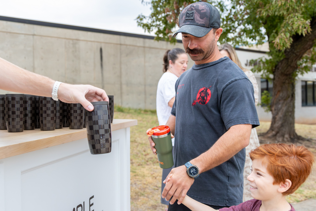 A father receives a black Simple Modern tumbler while his young son smiles beside him at the outdoor giveaway station.