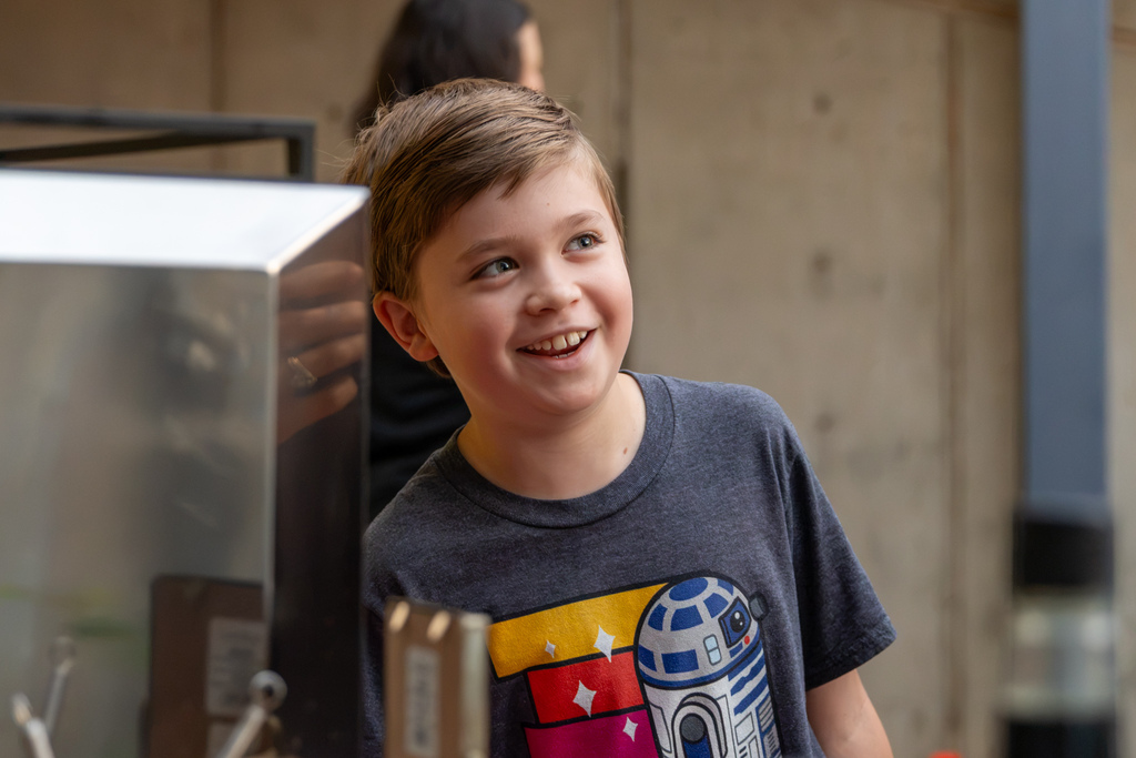 A smiling young student wearing a Star Wars T-shirt looks toward someone at the coffee station during the National Coffee Day celebration.